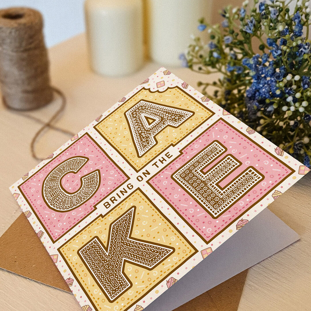 Pink and yellow birthday card with 'Bring On The Cake' text on a white wooden dresser with brown twine and blue flowers.