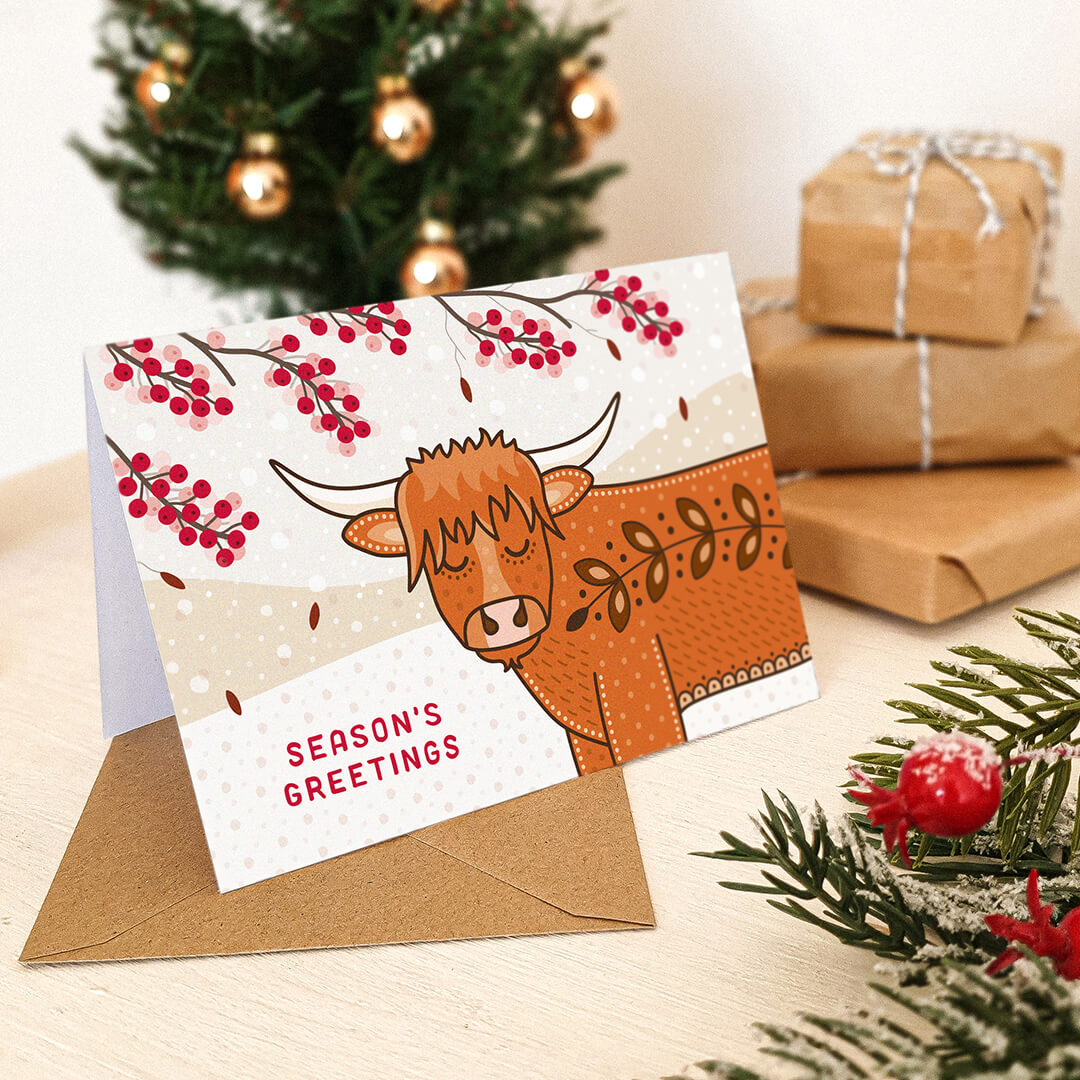 A festive greeting card featuring a cute Highland cow illustration with red berries and the words 'SEASON'S GREETINGS,' displayed with a brown kraft envelope on a tabletop with wrapped gifts and a Christmas tree in the background.