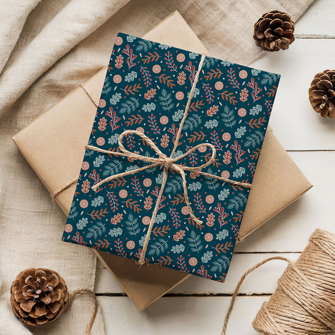 Gift wrapped in dark blue botanical Christmas wrapping paper with brown string, surrounded by pine cones on a wooden surface.