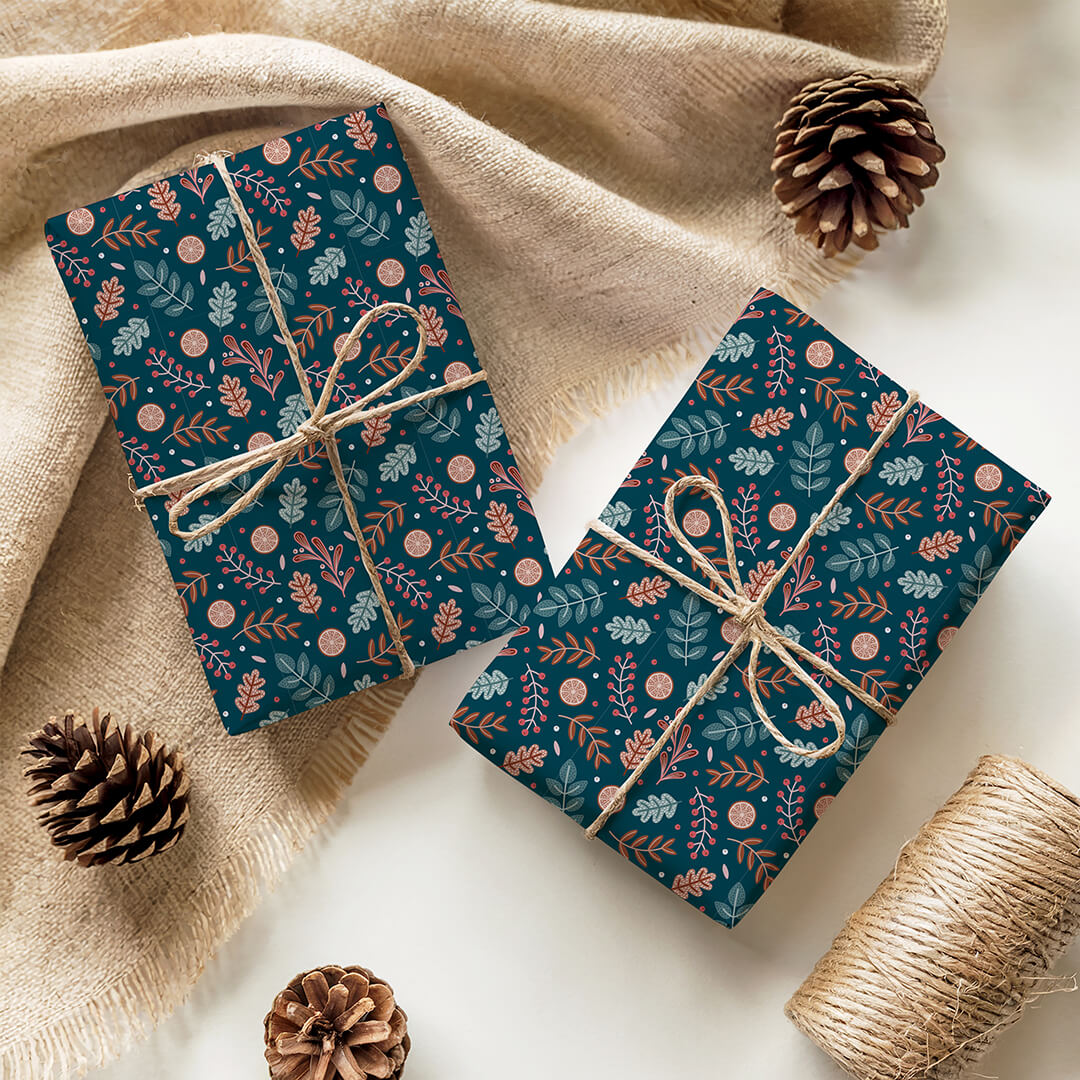 Two presents wrapped in botanical Christmas gift wrap and string, surrounded by pine cones and twine on a beige fabric background.