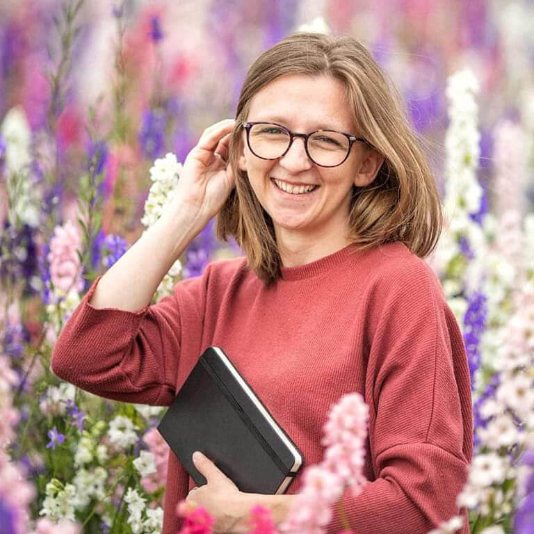 Jess, the owner of Jess A Little Creative, wearing a red jumper, smiling at camera holding a black notebook, stood in a field of pink and purple wildflowers