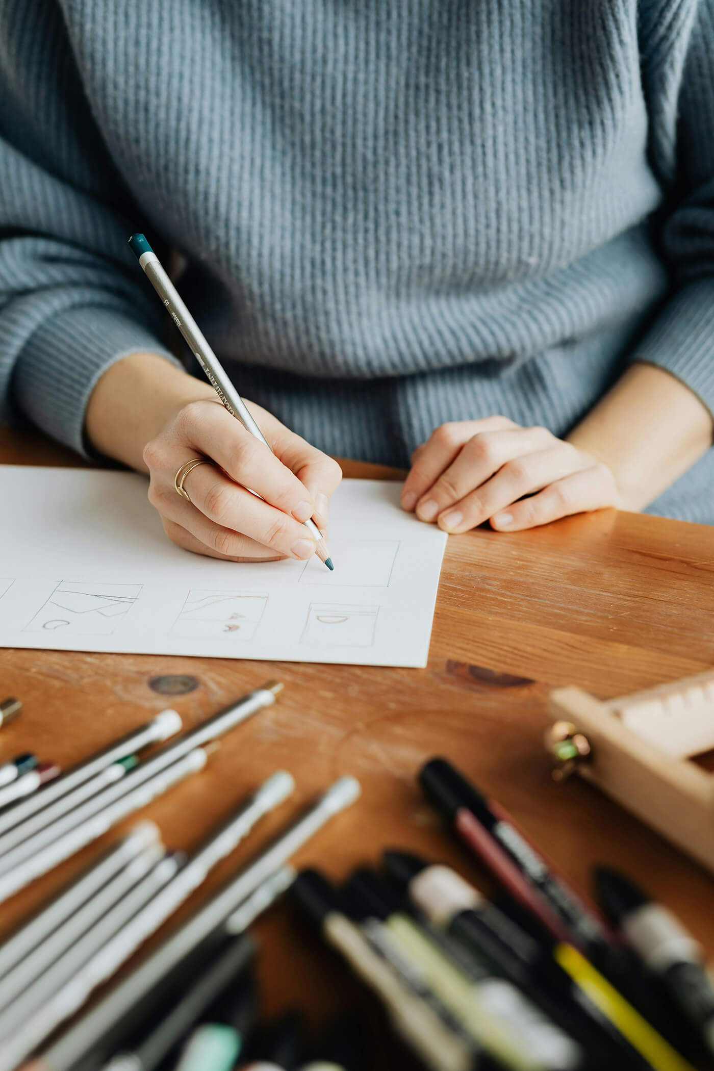 woman wearing grey jumper sat at a wooden desk sketching ideas onto paper surrounded by art materials