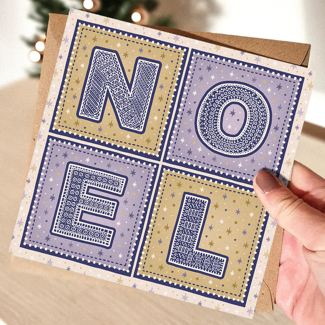 Purple and gold Noel Christmas card with 'NOEL' text held by a hand, Christmas tree in the background