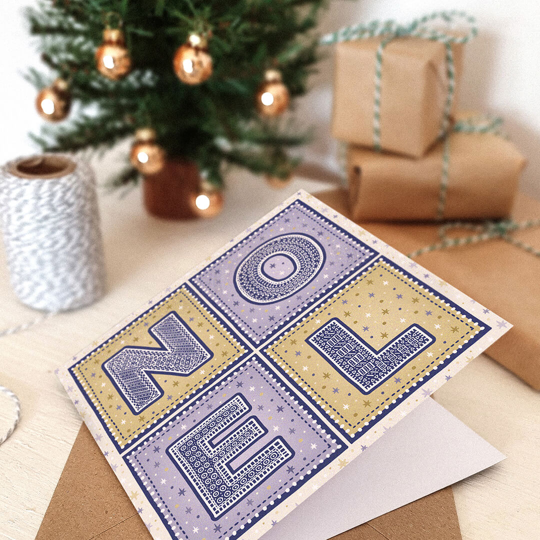 Purple and gold Noel Christmas card with 'NOEL' text in a festive setting with wrapped gifts and a small Christmas tree.