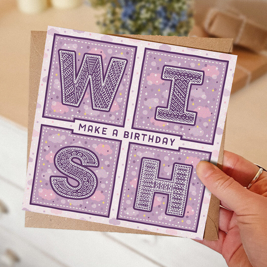 Hand holding a purple birthday card with 'Make a Birthday Wish' text on a blurred background