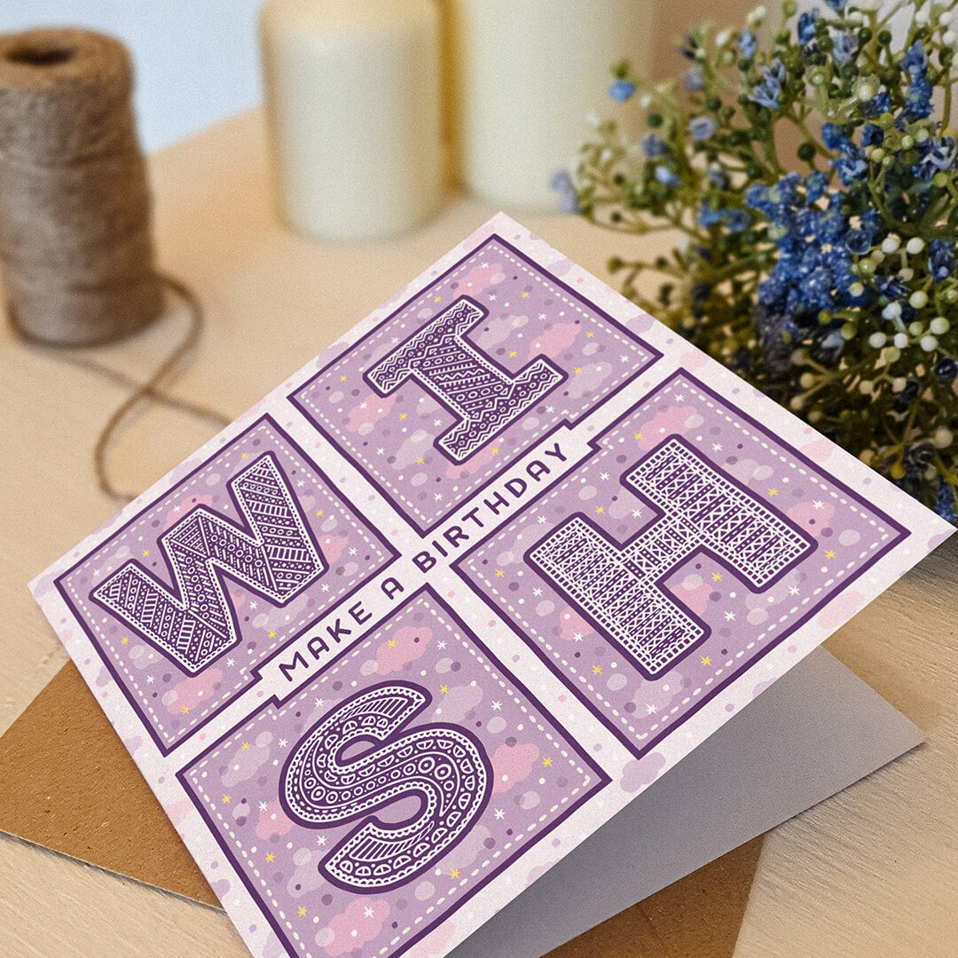Purple birthday card with decorative text spelling 'Make A Birthday Wish' on a white wooden dresser with a plant and candles in the background.
