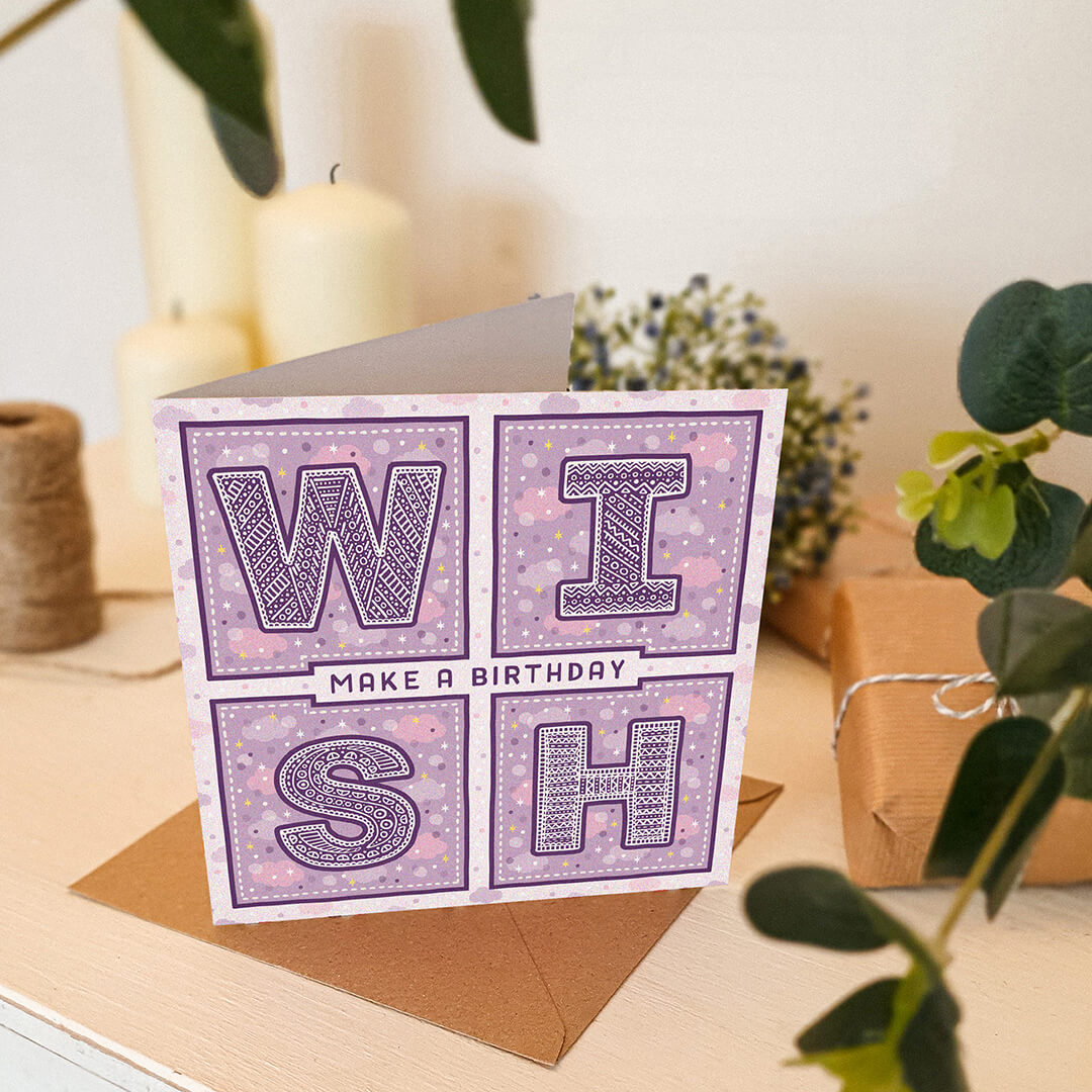 Purple birthday card with 'Make a Birthday Wish' text sat on a white wooden dresser with plants and candles in the background