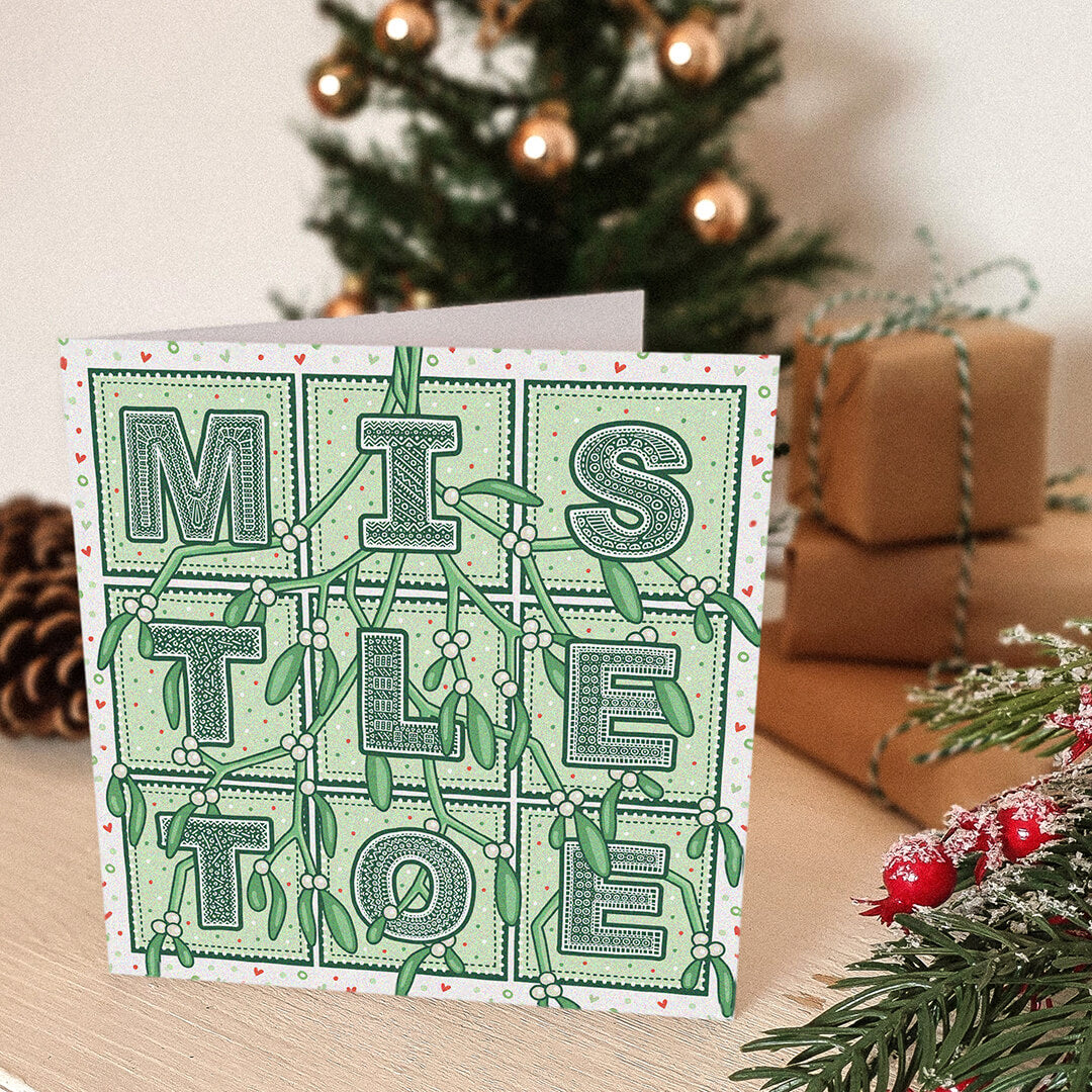 Romantic Mistletoe Christmas card with 'Mistletoe' typography and mistletoe illustrations in front of a decorated Christmas tree and presents.