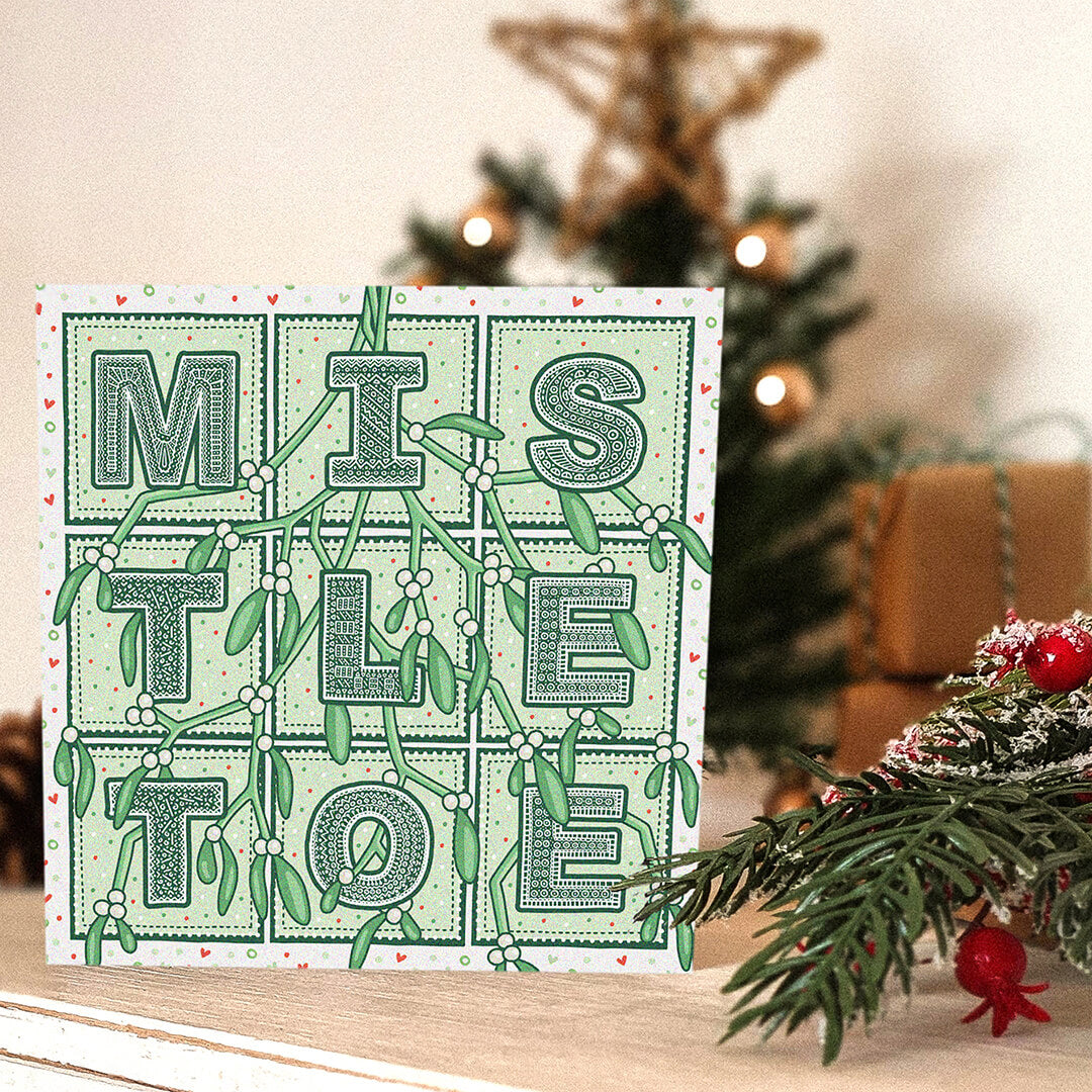 Romantic Mistletoe Christmas card with 'Mistletoe' typography and mistletoe illustrations, placed on a white wooden dresser with a blurred Christmas tree in the background.