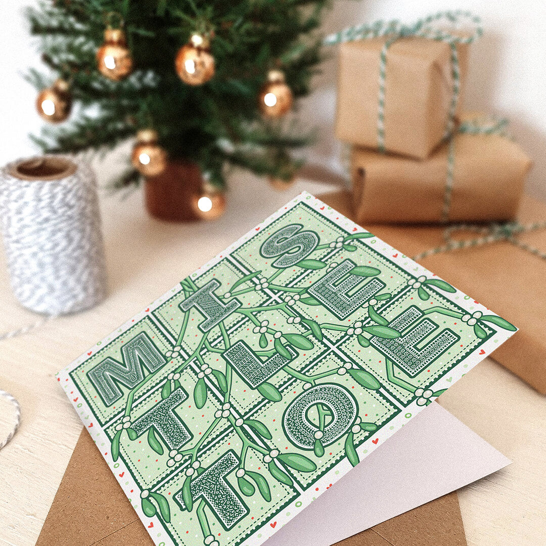 Romantic Mistletoe Christmas card with mistletoe typography and mistletoe illustrations on a white wooden dresser with wrapped gifts and a Christmas tree in the background