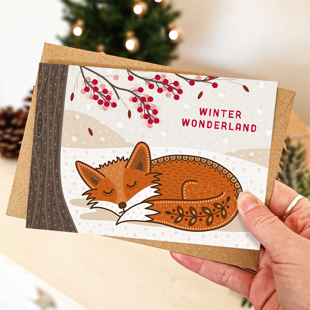 Hand holding snowy winter fox Christmas card with blurred festive background