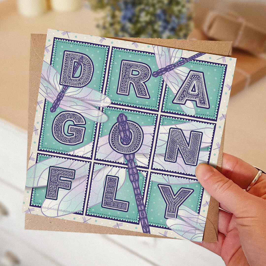 Purple and turquoise dragonfly greeting card with 'Dragonfly' text and dragonfly illustrations held by a hand on a blurred background