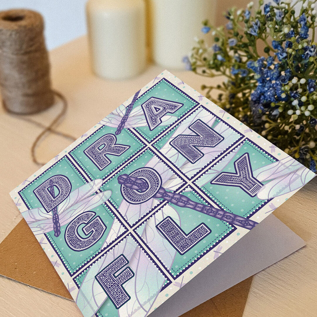 Purple and turquoise dragonfly greeting card with dragonfly illustrations and typography on a white wooden dresser with plants and candles in the background.