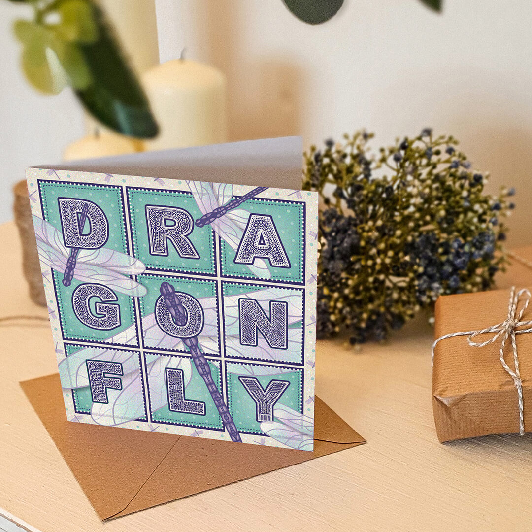 Purple and turquoise dragonfly greeting card with 'Dragonfly' text and dragonfly illustrations on a white wooden dresser with candles and plants in the background.