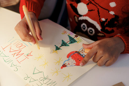 Young child sits at a desk writing a Christmas card wearing a red Christmas jumper