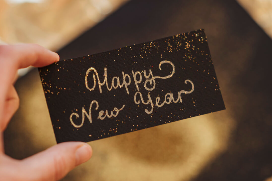 hand holding a small black card with 'Happy New Year' written in curly gold glittery lettering