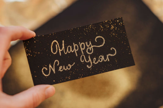 hand holding a small black card with 'Happy New Year' written in curly gold glittery lettering
