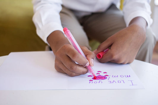 Child leant over a white table writing inside a Mother's Day card with a pink pen