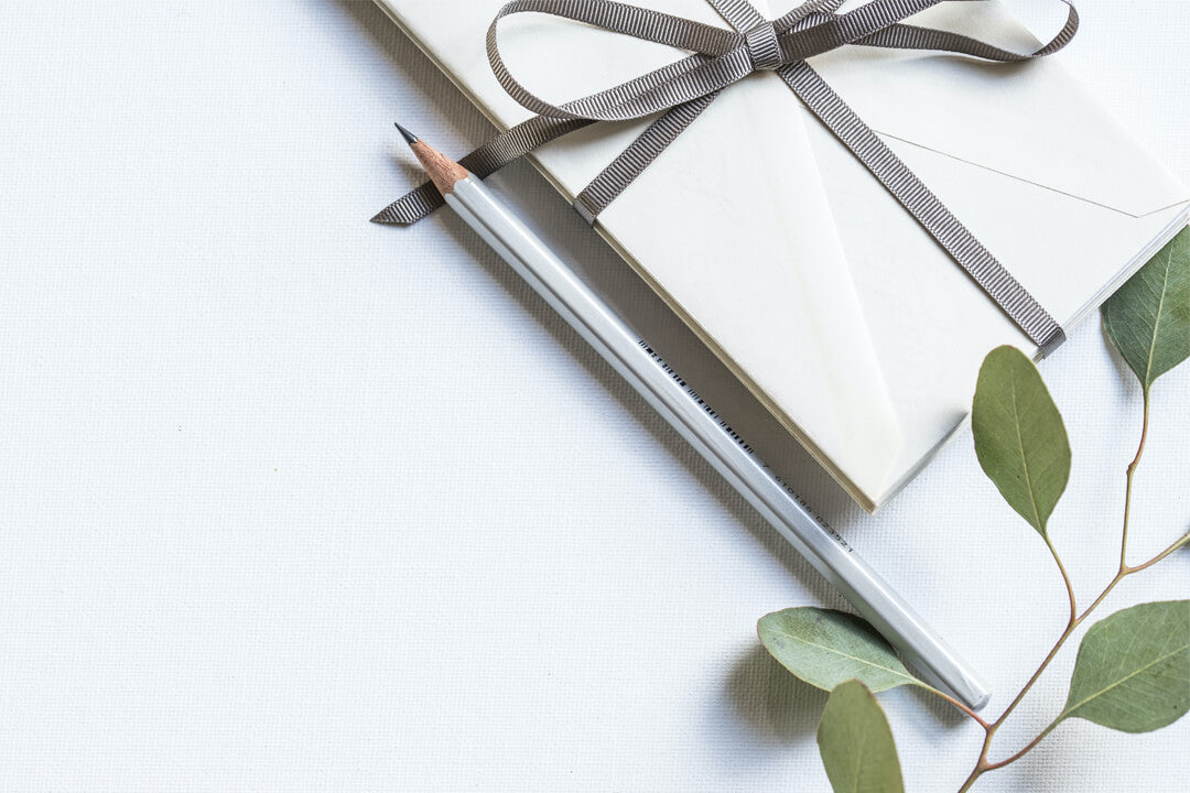 Pile of white wedding envelopes tied with silver ribbon, a silver pencil, and a eucalyptus branch all sit on an elegant white wedding table