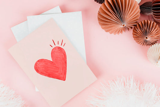 light pink valentine's day card with red heart and white envelope laid flat on a baby pink background with props