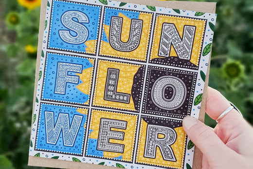 Sunflower illustration charity greeting card, held up in front of a field of sunflowers at sunset