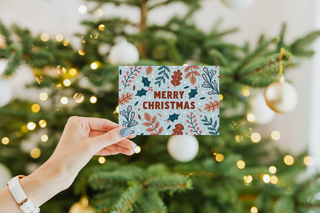 Festive foliage botanical Christmas card held up in front of a Christmas tree with gold lights and cream baubles