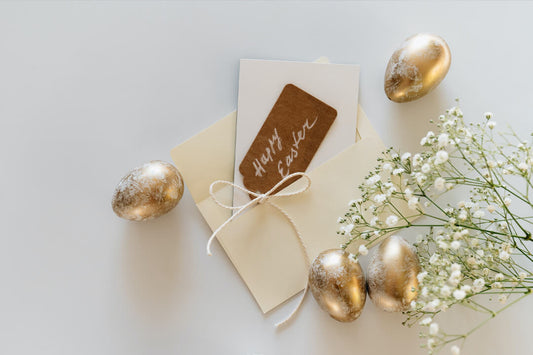 cream envelope and brown gift tag, sat on a white surface surrounded by small white flowers and golden easter eggs
