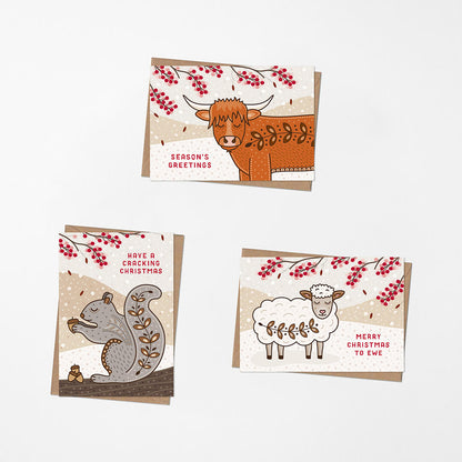 Three illustrated Christmas cards arranged on a light background. The cards feature animals and red berry branches: a highland cow with the text 'Season's Greetings,' a grey squirrel with 'Have a Cracking Christmas,' and a white sheep with 'Merry Christmas to Ewe'.