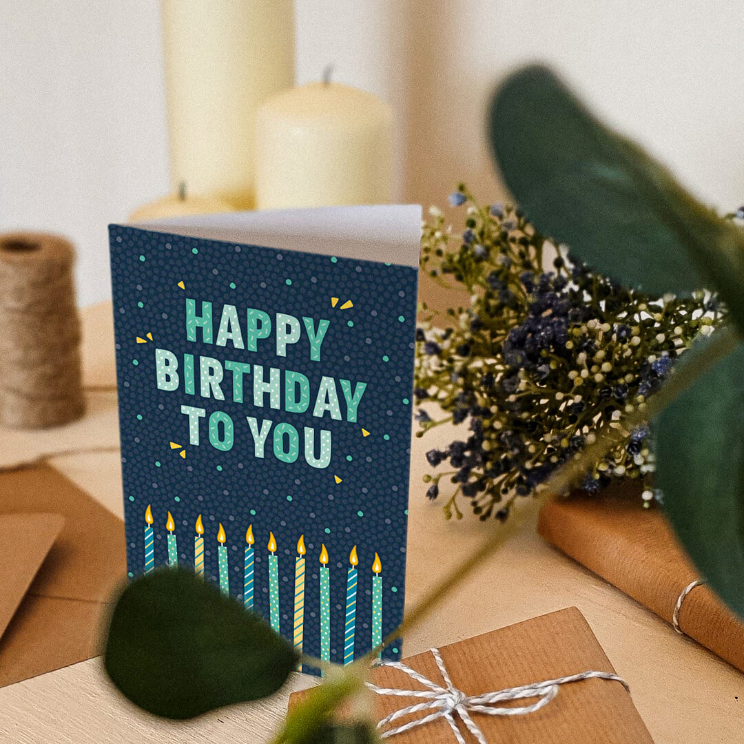 Blue and green happy birthday card sat on a shelf with a kraft brown envelope, brown twine, candles, flowers, and gifts wrapped in brown paper