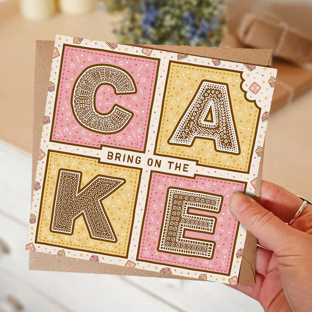 Pink and yellow birthday card with 'Bring On The Cake' text held by a hand, with a blurred background