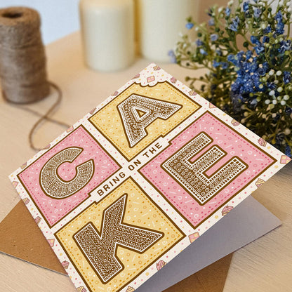 Pink and yellow birthday card with 'Bring On The Cake' text on a white wooden dresser with brown twine and blue flowers.