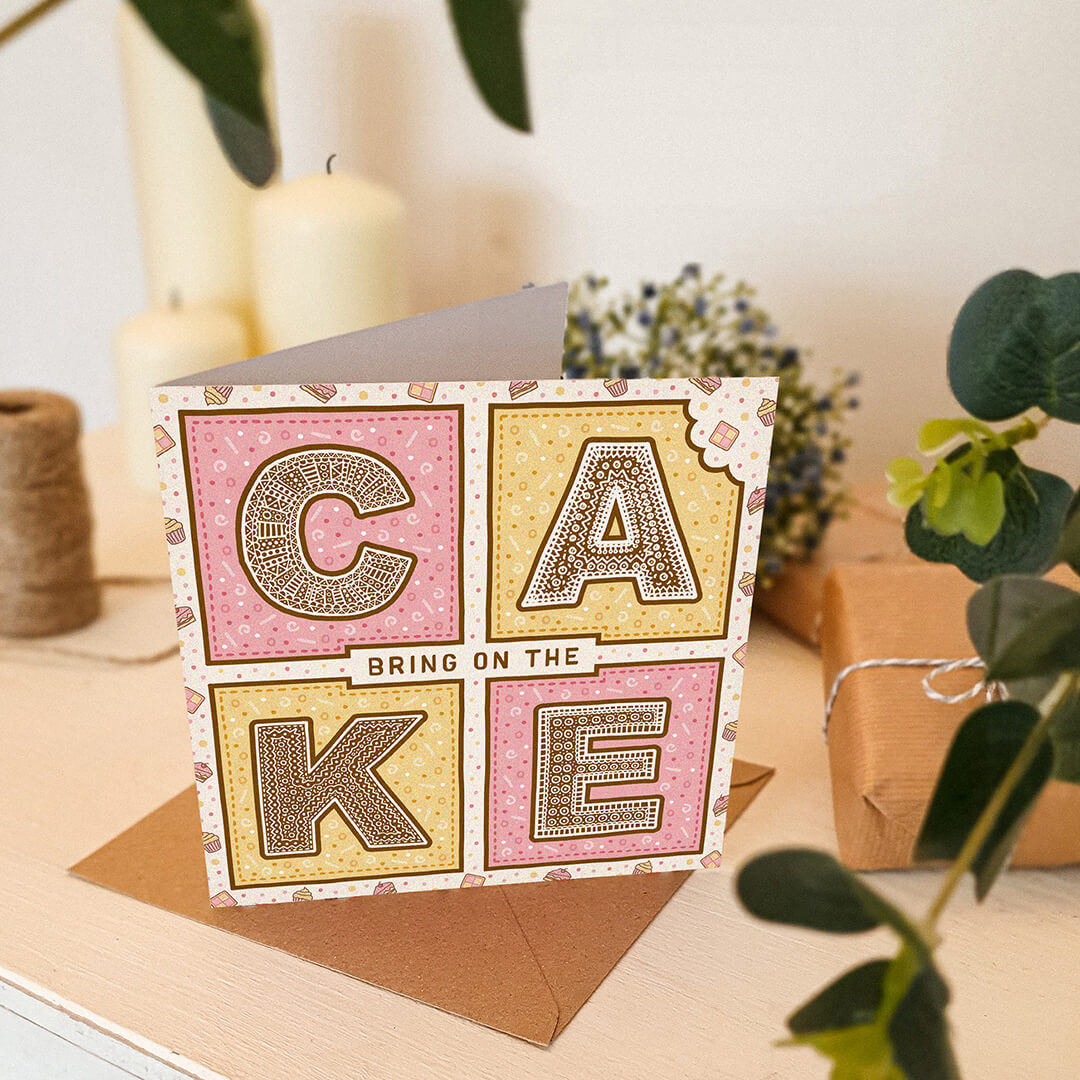 Pink and yellow birthday card with 'Bring On The Cake' text on a white wooden dresser with plants and candles in the background