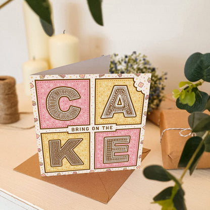 Pink and yellow birthday card with 'Bring On The Cake' text on a white wooden dresser with plants and candles in the background