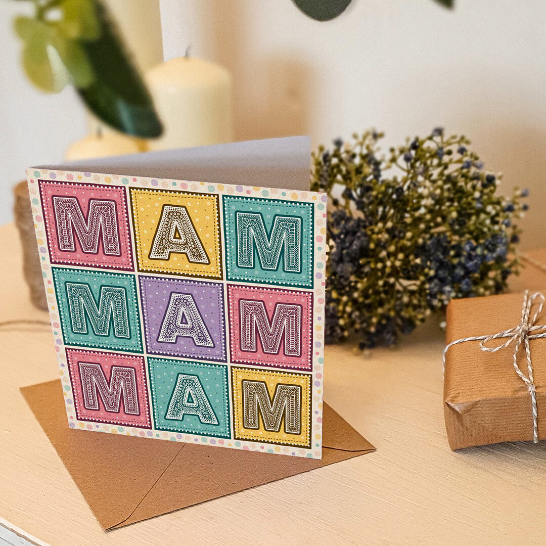 Colourful Mother's Day card with 'MAM' message on a cream wooden table with gifts and flowers.