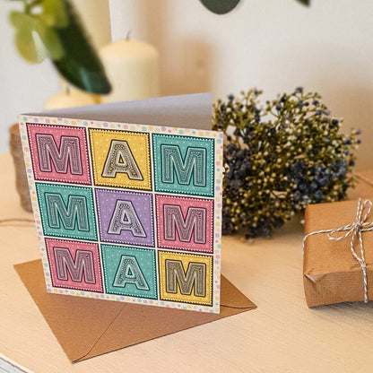 Colourful Mother's Day card with 'MAM' message on a cream wooden table with gifts and flowers.