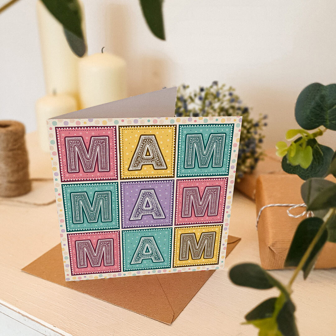 Colourful Mother's Day card with 'MAM' message on a cream wooden table with plants and candles in the background