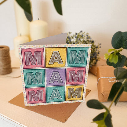 Colourful Mother's Day card with 'MAM' message on a cream wooden table with plants and candles in the background