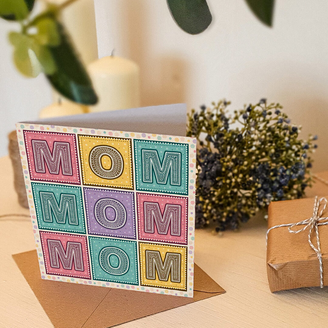 Mother's Day card with 'MOM' message on a cream wooden table with a plant and candles in the background