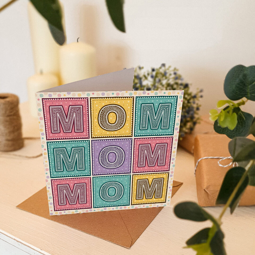Colourful 'Mom' Mother's Day card on a cream wooden table with plants and candles in the background