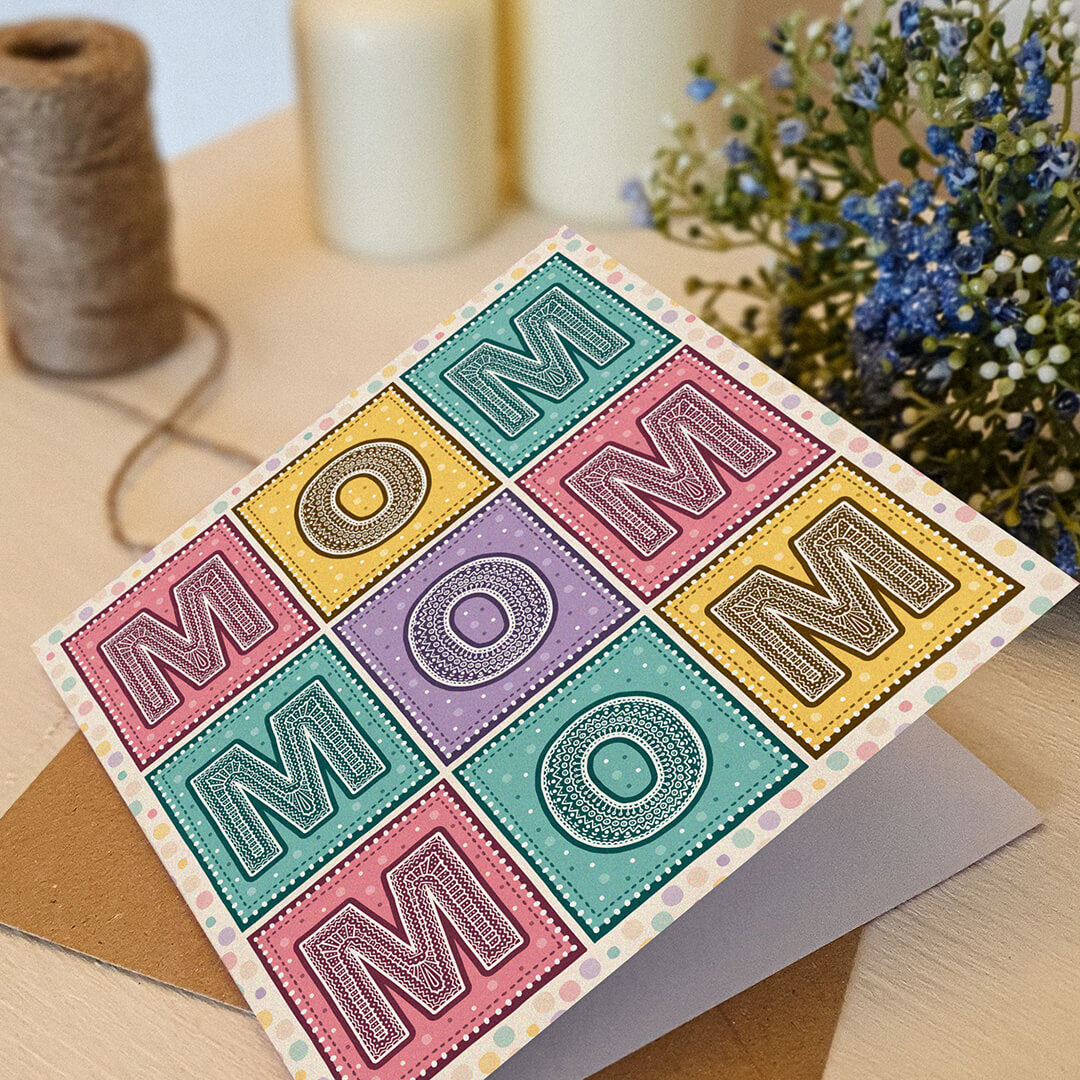 Colourful Mother's Day card with 'MOM' message on a cream wooden table with decorative items.