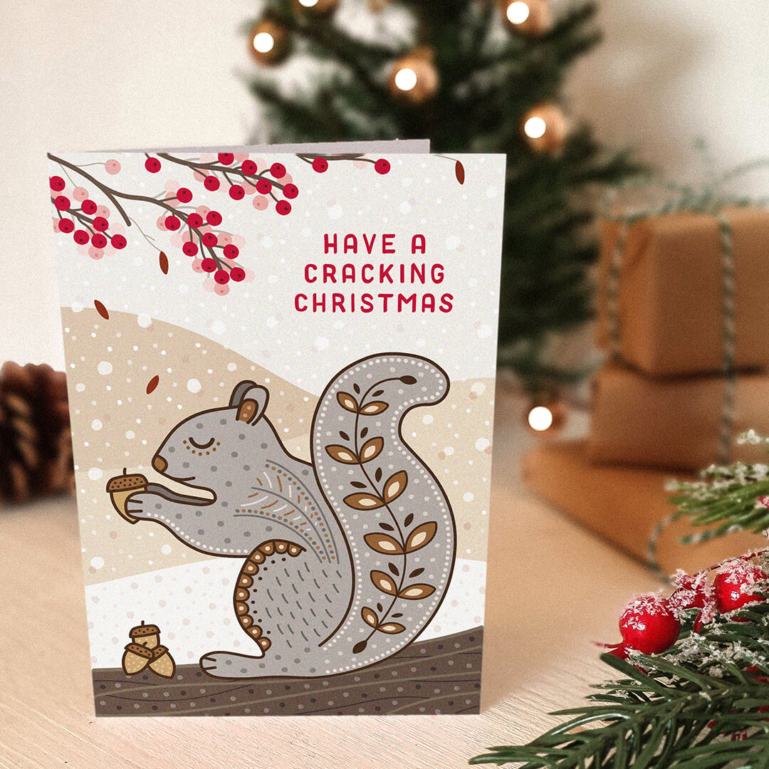 cute illustrated grey squirrel christmas card, sat on a tabletop with a blurred christmas tree and gifts in the background.