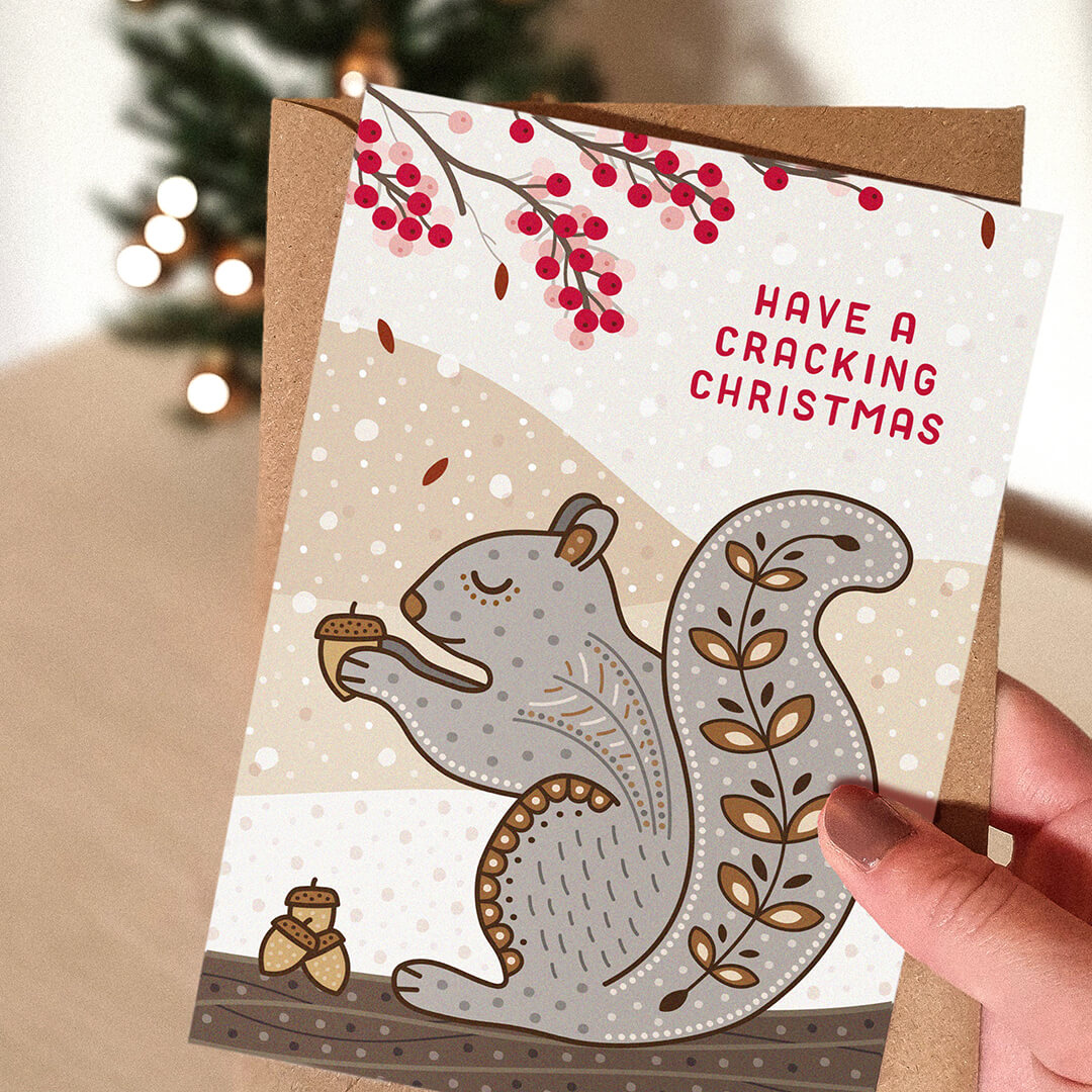 hand holding a grey squirrel christmas card design with kraft brown envelope with blurred christmas tree in the background.