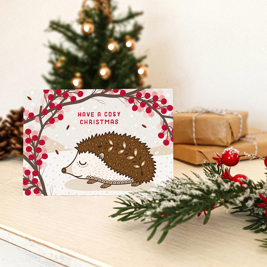 Unique Christmas card with hedgehog illustration and festive ornaments on a cream wooden table.