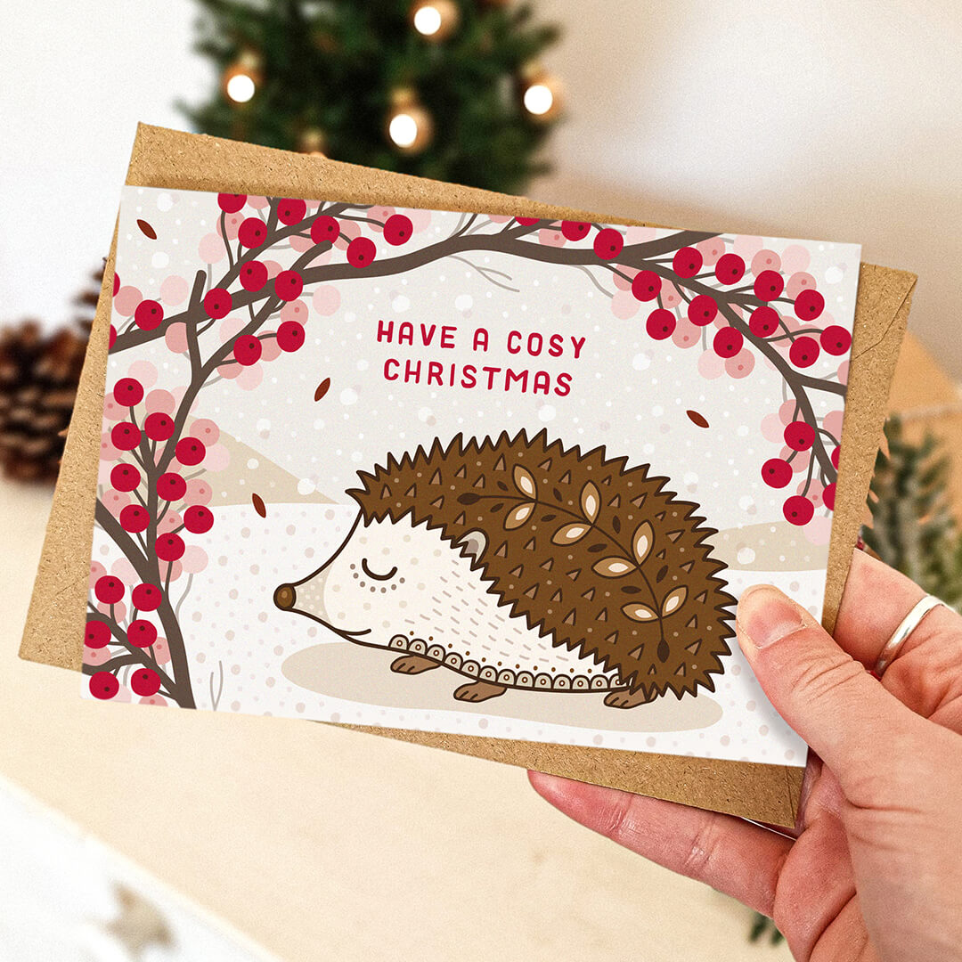 Hedgehog Christmas card with 'Have a Cosy Christmas' message held by a hand, with Christmas tree blurred in background.
