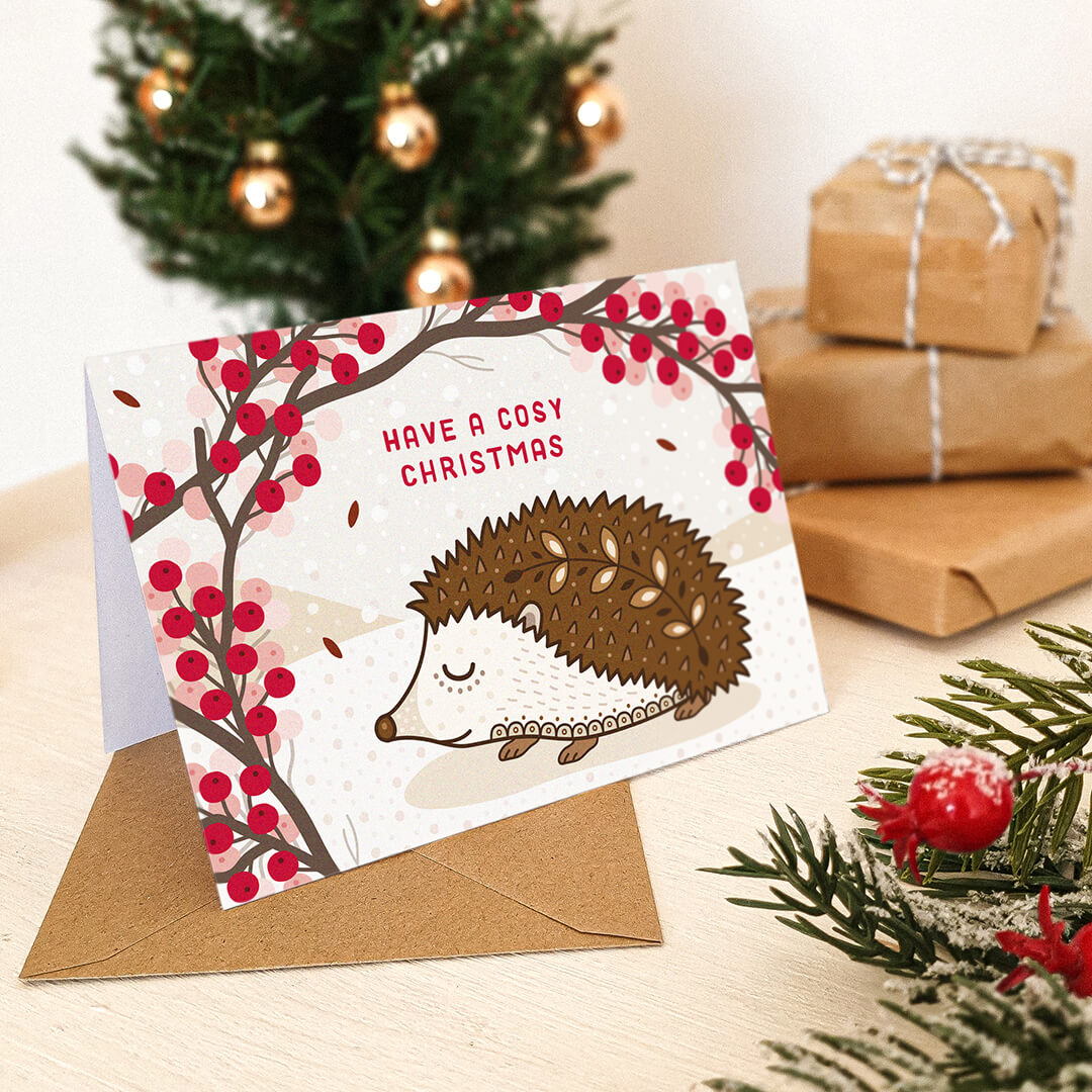 Christmas card with hedgehog design and 'Have a Cosy Christmas' message, on a cream wooden table with festive decorations.