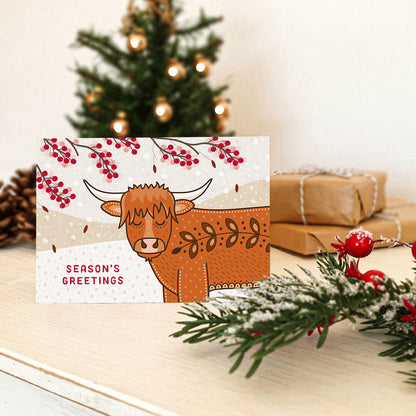 A Christmas card featuring a cute Highland cow illustration, placed on a tabletop with a blurred Christmas tree, wrapped gifts, and holiday greenery in the background.