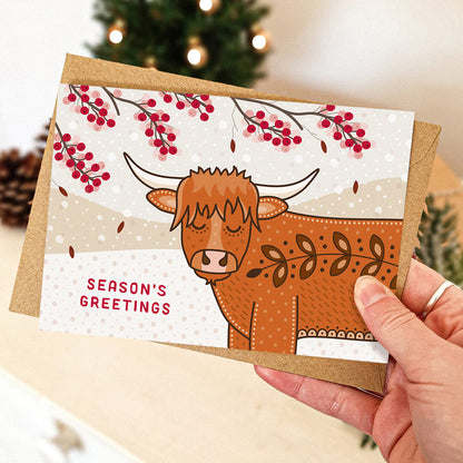 A hand holds a Christmas card featuring a cute Highland cow illustration, red berry branches, and the text 'SEASON'S GREETINGS' with a brown kraft envelope, with a softly blurred holiday background.