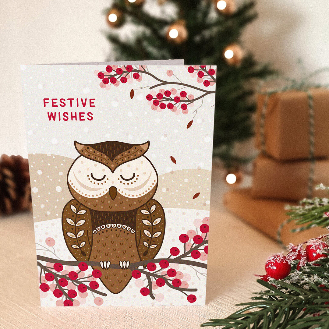 Christmas card with owl design and 'Festive Wishes' message, placed on a cream wooden table with festive decorations.