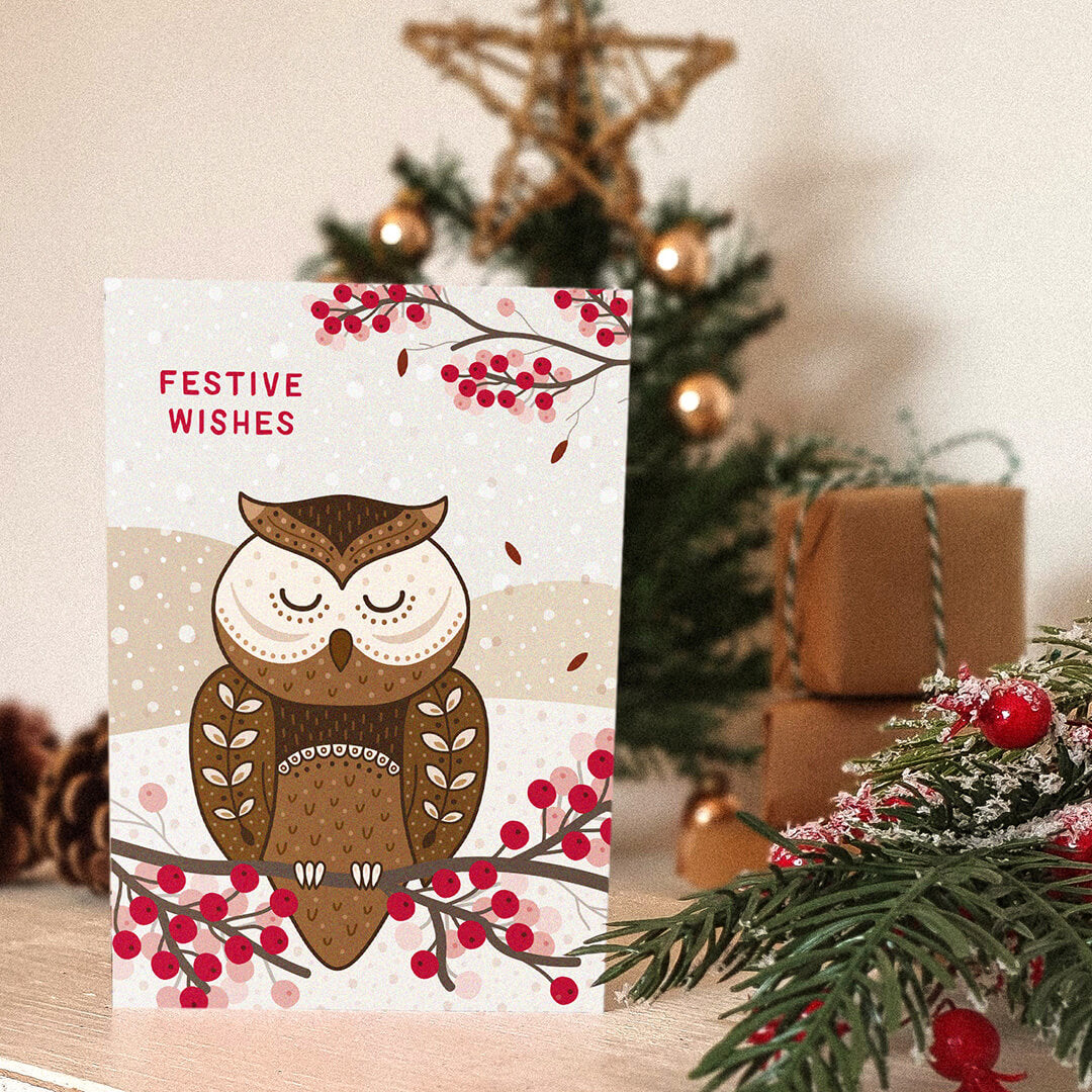 Christmas card with owl design and 'Festive Wishes' message, placed in front of a decorated Christmas tree and gifts.