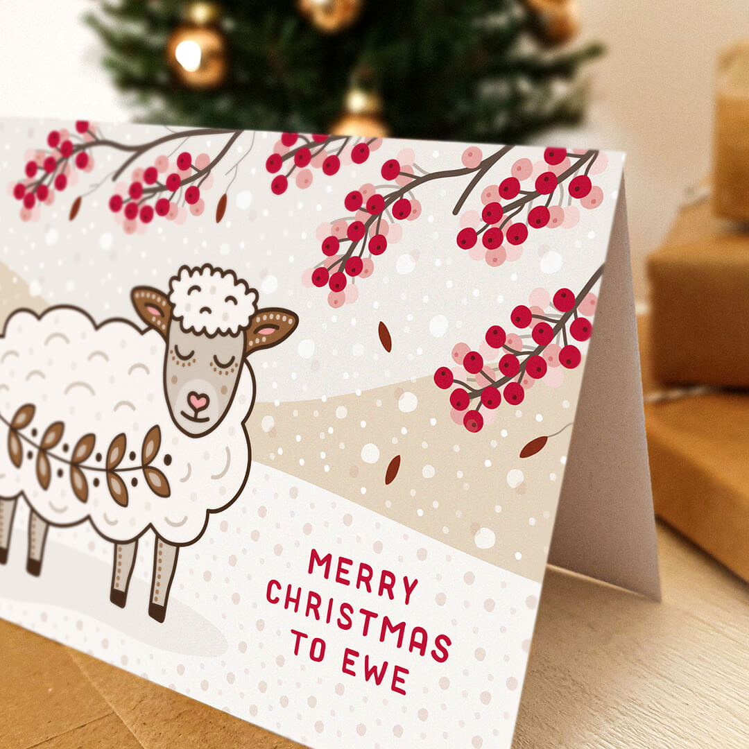 A cute sheep christmas card on a tabletop with blurred christmas tree and gifts in the background